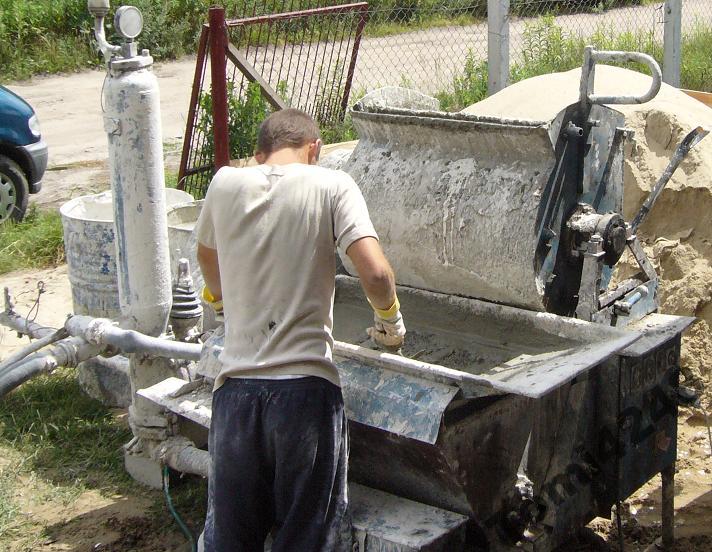 Pracownik w trakcie mieszania tynku cementowo-wapiennego z piaskiem kwarcowym w dużej mieszarce budowlanej na zewnątrz, widoczny zbiornik ciśnieniowy i stos piasku.